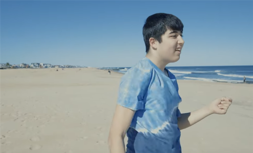 A picture of the student in the video clip on a sandy beach with houses and waves in the background.